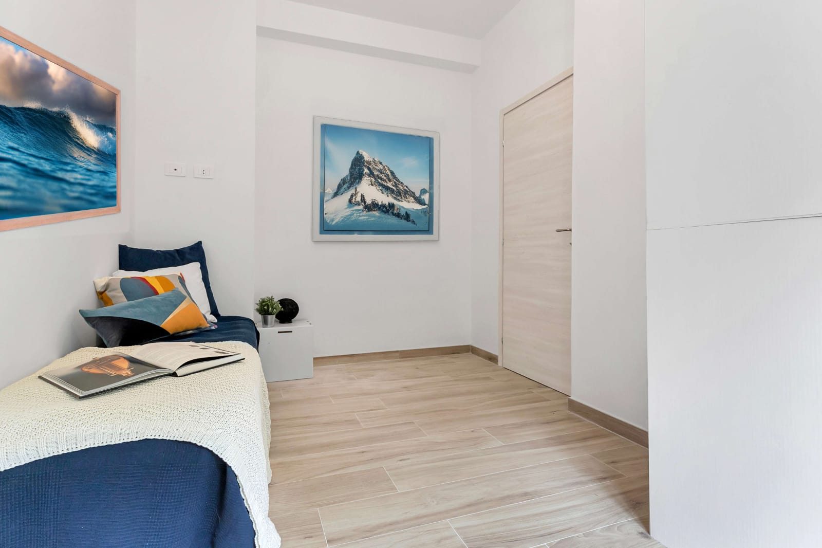Camera singola con letto, due quadri alle pareti, porta interna chiara e pavimento effetto legno