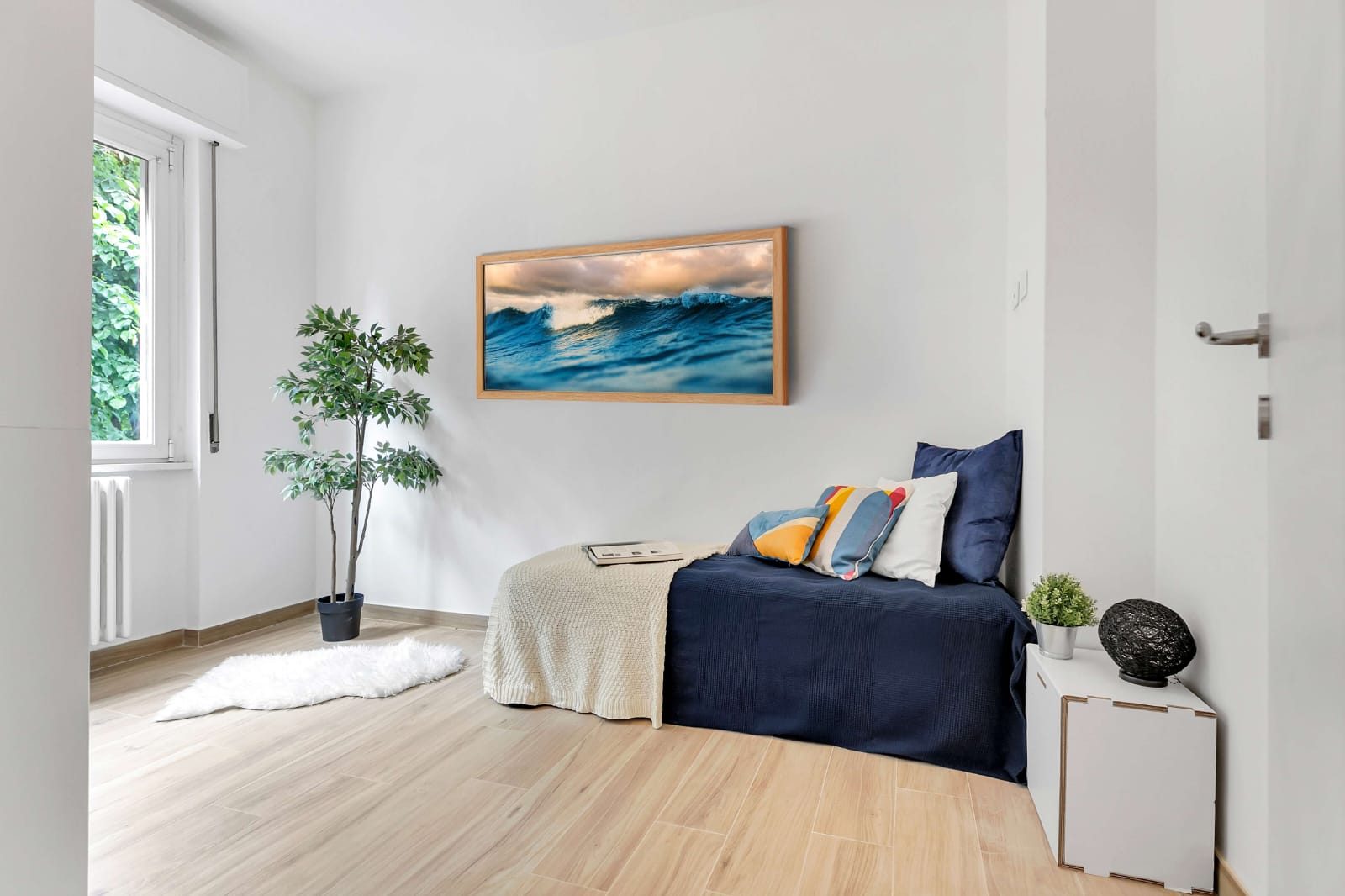 Camera singola con letto e copriletto blu, quadro alla parete, pianta e finestra con radiatore