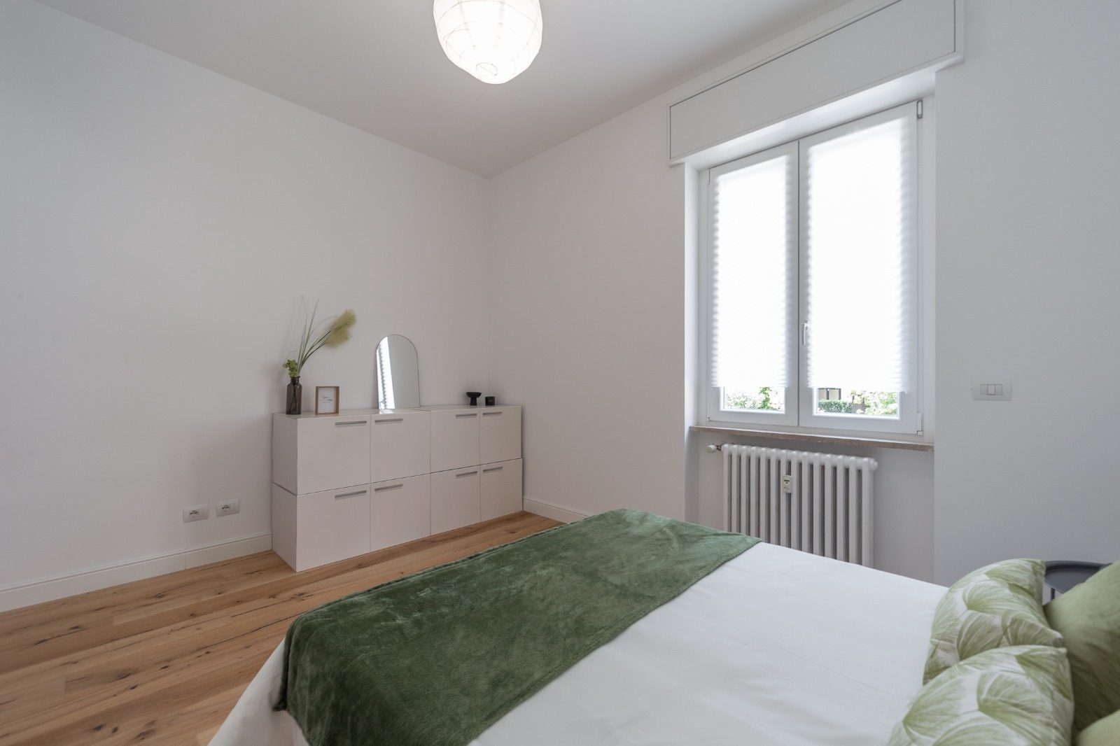 Camera matrimoniale con parquet in rovere, finestra con tenda plissettata e radiatore a colonne