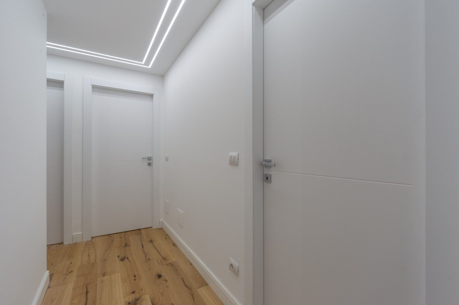 Corridoio con pareti bianche, porte interne bianche, pavimento in legno e profilo LED a soffitto
