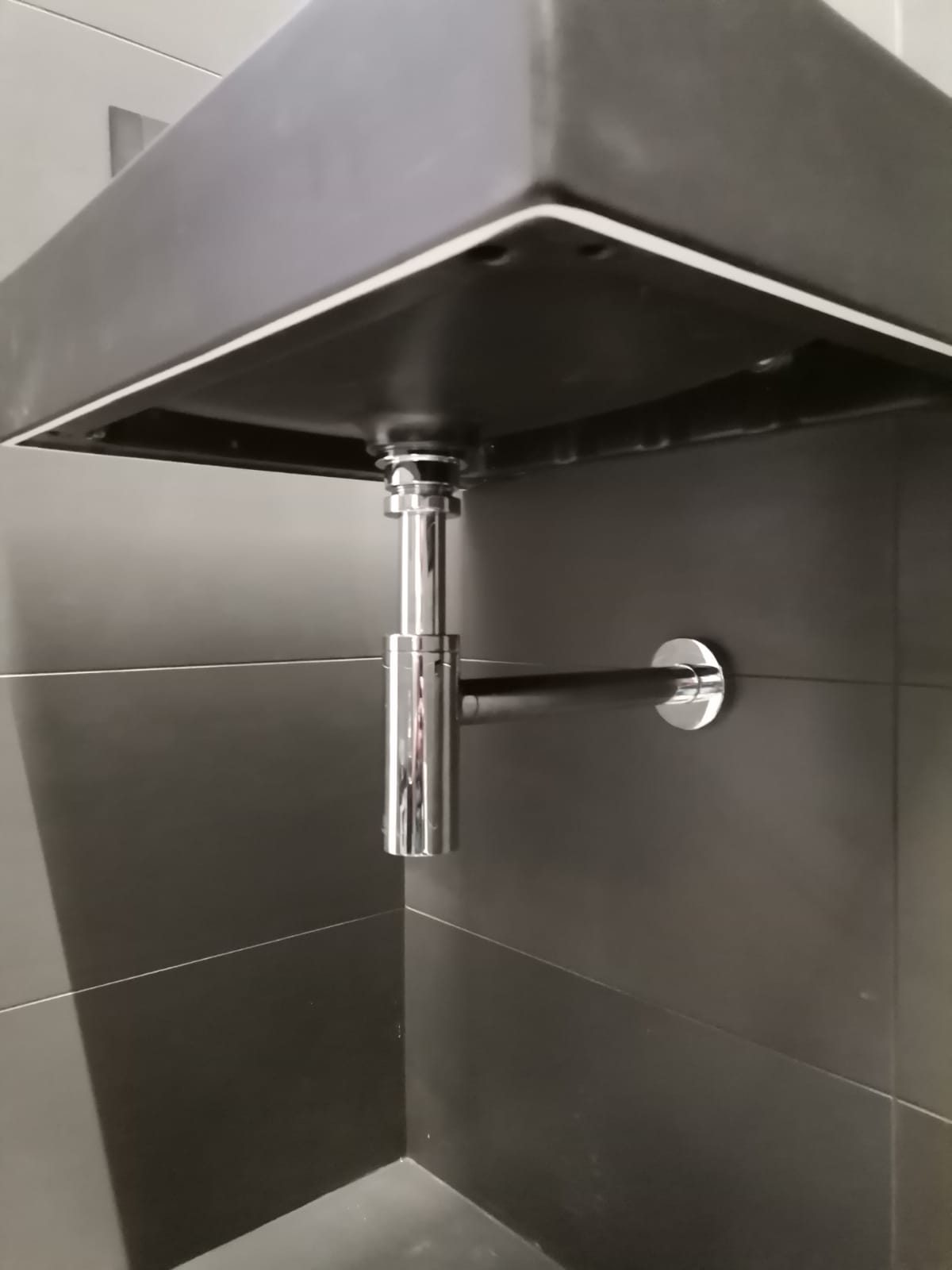 Piano lavabo scuro sospeso visto dal basso, con sifone cromato e scarico a parete