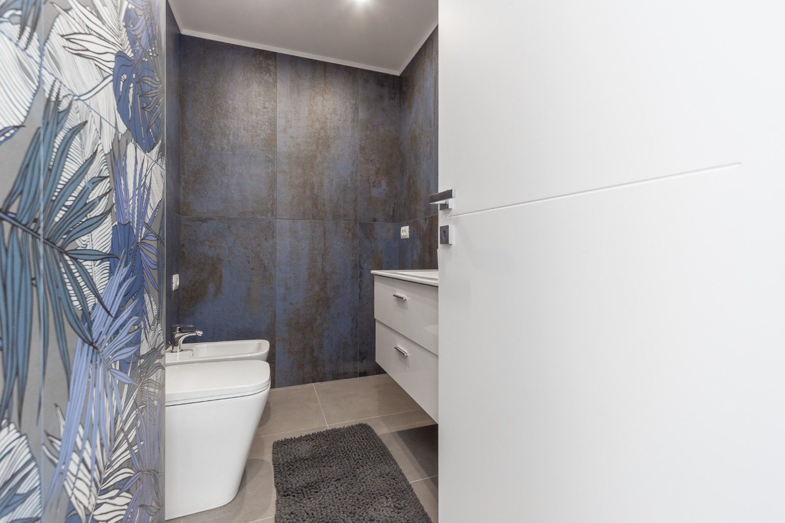 Bagno con grandi lastre effetto metallo, parete decorata a foglie, sanitari bianchi e mobile lavabo