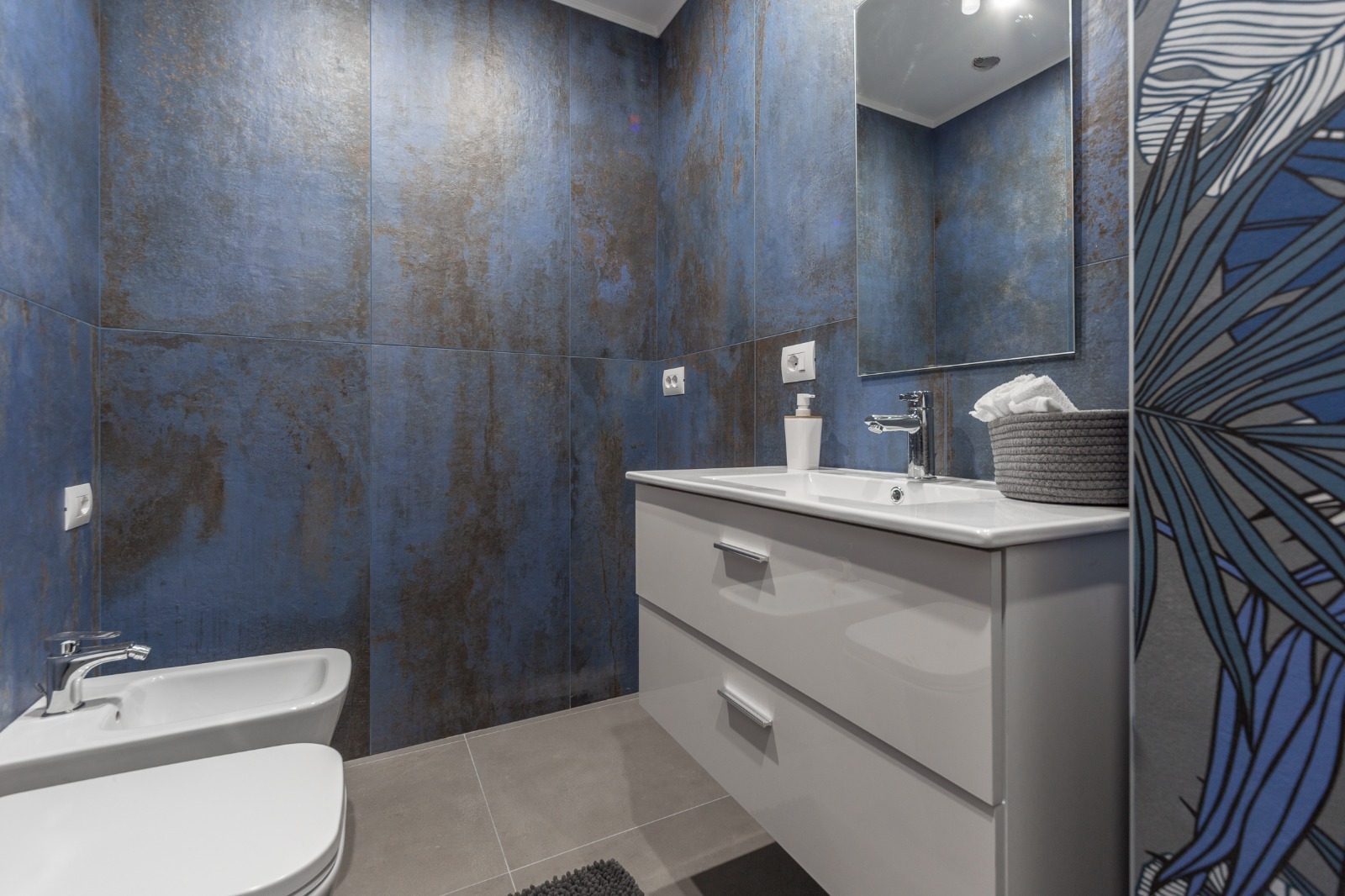 Bagno con rivestimento blu effetto metallo ossidato, mobile sospeso bianco, lavabo, bidet e specchio