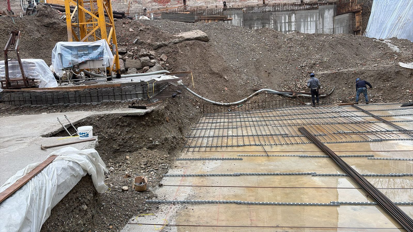 Cantiere con gru, pompa per calcestruzzo, rete di armatura stesa sul magrone e due operai