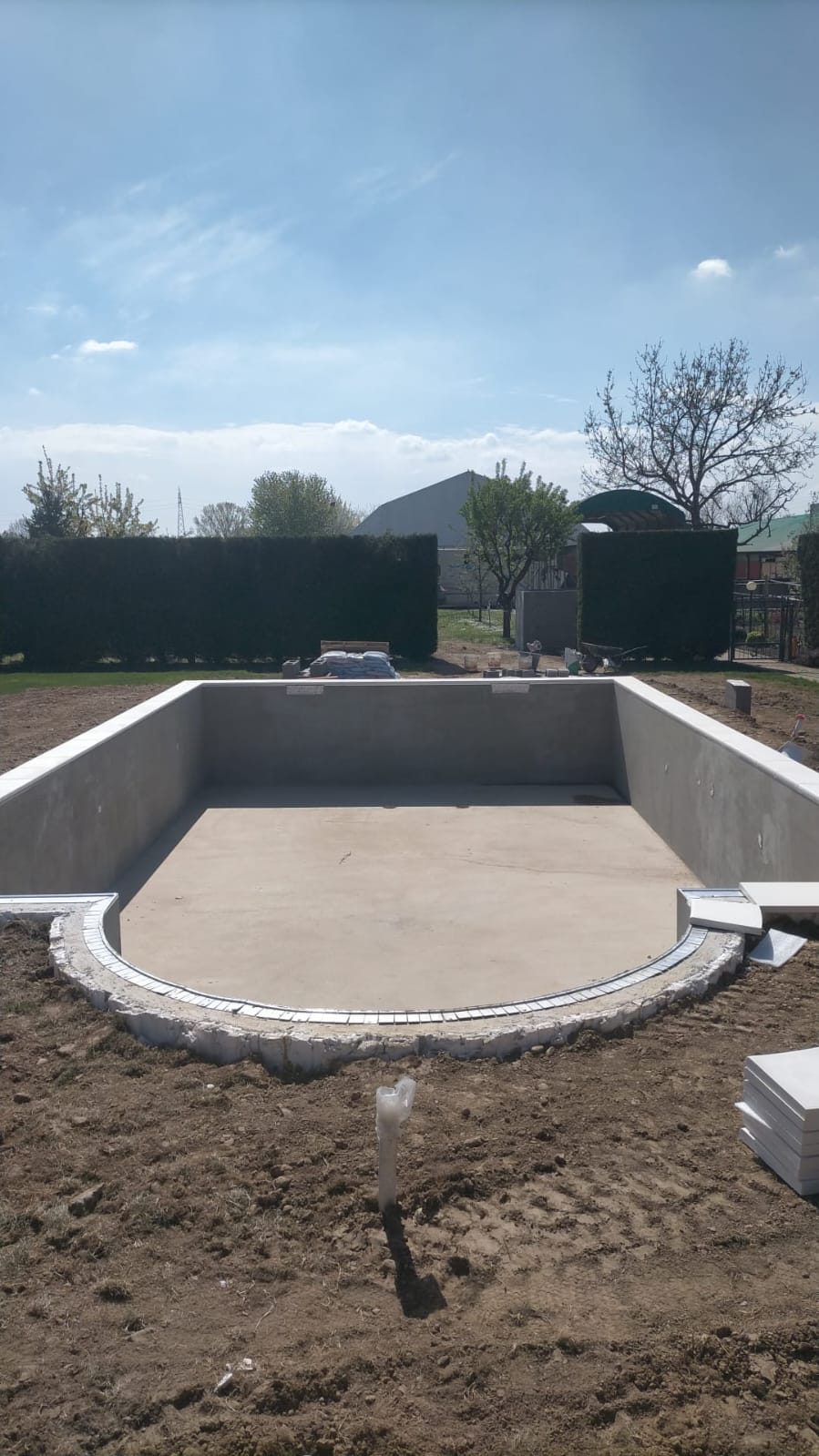 Vasca di piscina interrata in cemento intonacato, vuota, con bordo curvo e terreno scavato attorno.