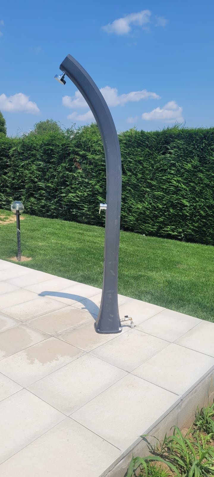 Colonna doccia solare da esterno installata sulla pavimentazione, con siepe e prato sullo sfondo