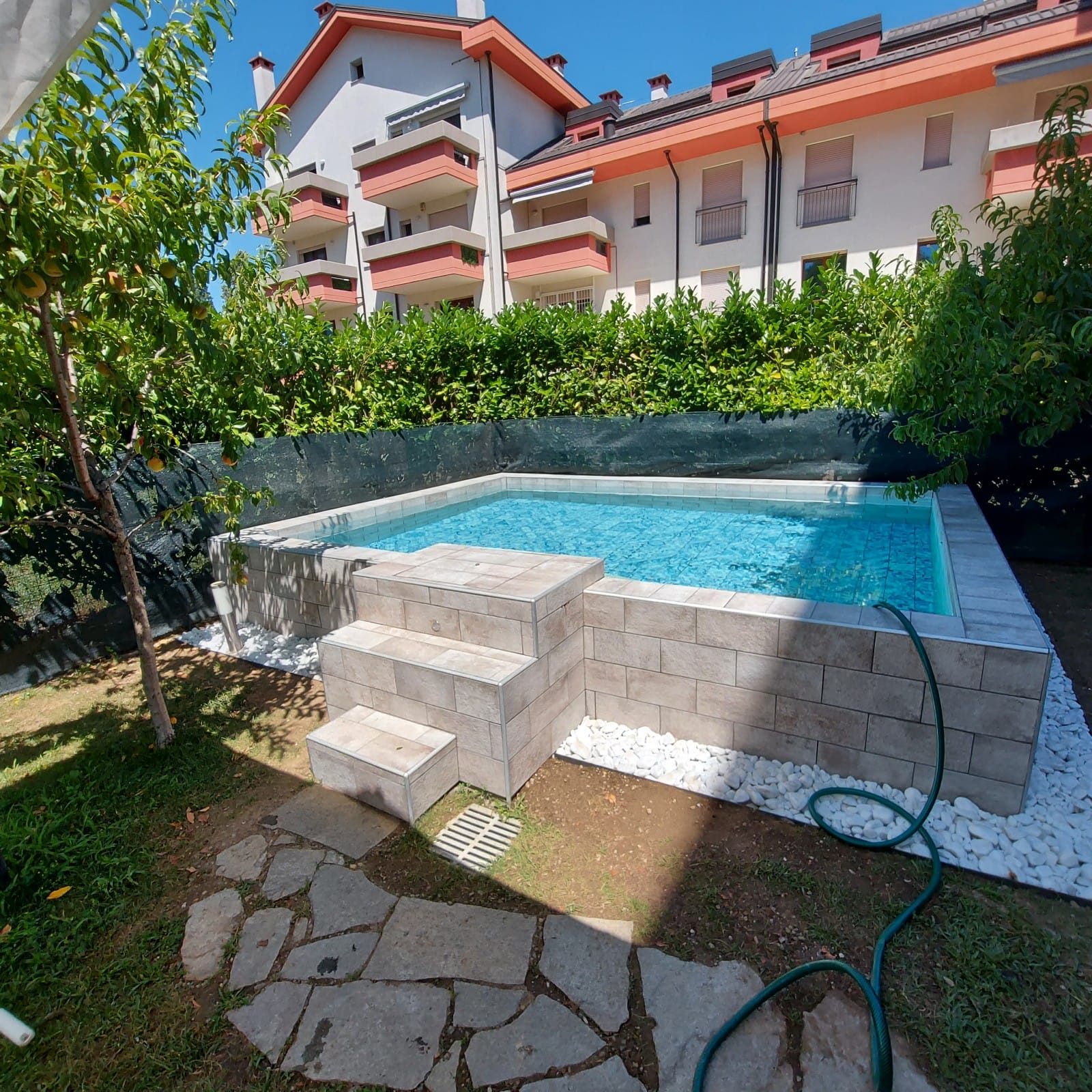 Piscina fuori terra rivestita in piastrelle effetto pietra, gradoni d'accesso e ciottoli bianchi