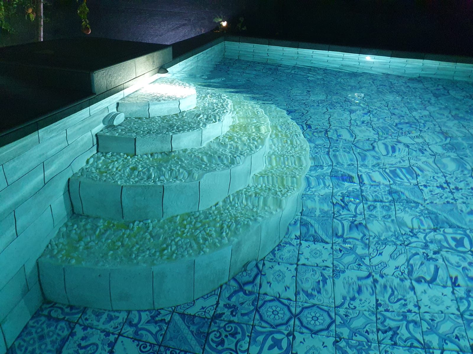 Piscina illuminata di notte con scala curva a gradoni e fondo rivestito in piastrelle decorate