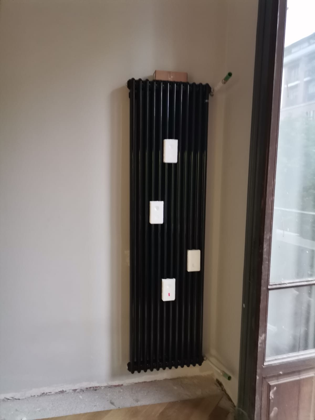 Radiatore tubolare verticale nero montato a parete accanto alla portafinestra, protezioni bianche ancora applicate